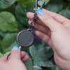 steel cable phone tether in black-gunmetal , view of front black leather patch and back, metal with logo embossed. 